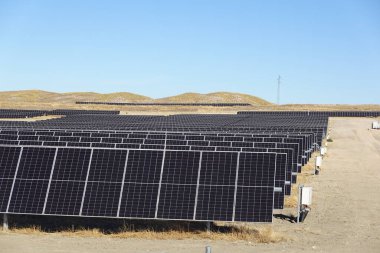 Büyük bir güneş çiftliğinde sıra sıra dizilmiş güneş panellerinin geniş görünümü, açık mavi gökyüzünün altında kurak tepeler var..