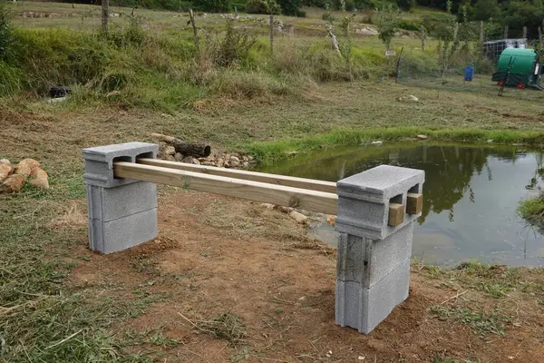 Beton bloklardan inşa edilmiş açık hava tezgahı ve kırsal bir çiftlikteki küçük bir göletin yanına yerleştirilmiş ahşap bir kalas. Barışçıl ortam, doğal elementlerle çevrili basit bir DIY projesini vurguluyor.