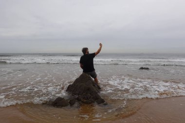 Okyanus kıyısındaki bir kayanın üzerinde oturan bir adam, etrafındaki dalgaların sakin ve bulutlu bir yaz gününde çarptığı bir selfie çekiyor.