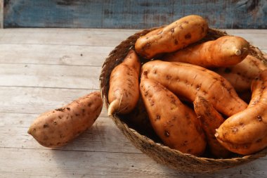 Birkaç tatlı patates ahşap bir masada hasır bir sepetin içinde oturuyor, dünyevi tonlarını ve dokularını sergiliyorlar.