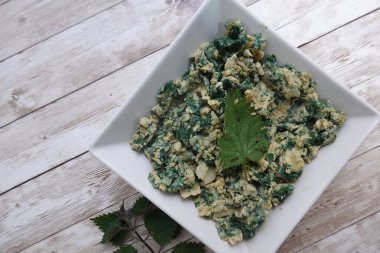 Isırgan otuyla çırpılmış yumurta ve soğan. Isırgan yaprağıyla süslenmiş. Tahta bir masada beyaz kare bir tabakta servis ediliyor.