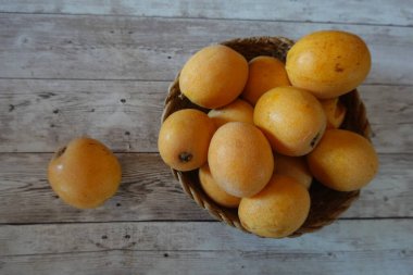 Olgun lokuatlar, Japon medyatörleri veya Japon erikleri olarak da bilinir, taşralı ahşap bir masa üzerinde sepetlenmiş bir hasır sepet taşarak canlı bir natürmort oluştururlar.