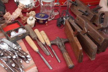 Ahşap işleme aletleri, çatal bıçak takımı ve eski incik boncuklar kırmızı bir masa örtüsünün üzerinde sergilenerek bitpazarının hazinelerini canlı bir şekilde sergiliyorlar.