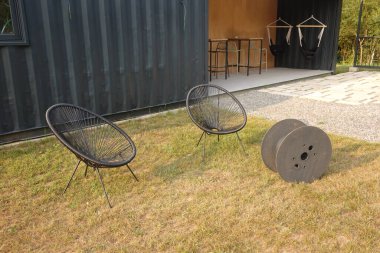 Two modern acapulco chairs and an empty cable reel standing on the lawn in front of a renovated shipping container house with hammocks and bar stools