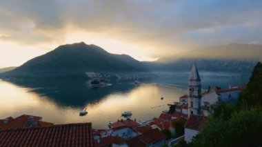 4K renkli hızlandırılmış görüntü Kotor Körfezi 'ndeki Perast kasabasının gün batımında renkli gökyüzü ışığı sızıntıları, dumanlar ve bulutlar ile dağların arkasında sakin deniz ve iki ikonik ada.