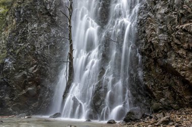 Şelale yosunlu kayaların arasından akıyor ve su uzun pozlama tekniğiyle bir peçe gibi çekiliyor.