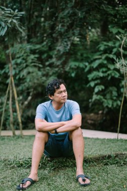 Güneydoğu Asyalı bir adam devrilmiş bir ağaç gövdesine oturmuş, boş boş uzaklara bakıyor, doğanın ortasında bir düşünce ve yalnızlık hissi uyandırıyor.