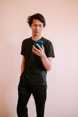 Güneydoğu Asyalı bir adam elinde akıllı bir telefon tutuyor ve dikkatle ekrana odaklanıyor. Gelişigüzel giyinmiş, modern ve teknoloji meraklısı bir yaşam tarzı sergiliyor, bağlantı ve iletişim temaları için mükemmel..