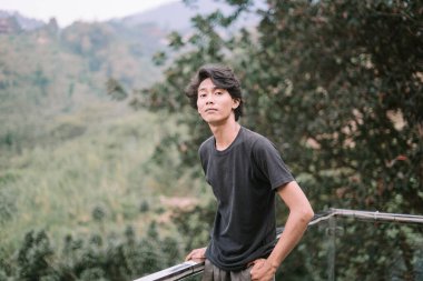 Güneydoğu Asyalı bir adam balkonda duruyor, yemyeşil ve huzurlu manzaranın tadını çıkarıyor.