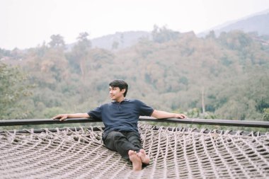 Güneydoğu Asyalı bir adam, arka planda nefes kesici bir manzarası olan hamak tarzı bir ağda dinleniyor, doğada huzurlu bir anın tadını çıkarıyor.