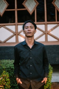 Siyah gömlek giyen Güneydoğu Asyalı bir adam kameranın önünde şık bir poz veriyor.