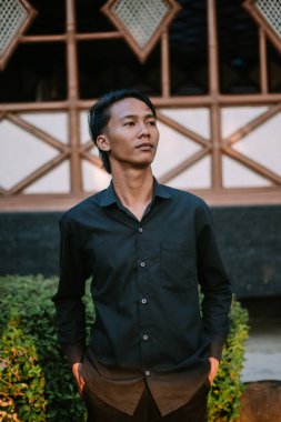 Siyah gömlek giyen Güneydoğu Asyalı bir adam kameranın önünde şık bir poz veriyor.