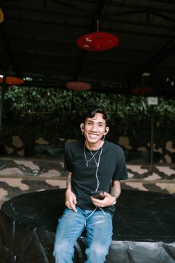 Güneydoğu Asyalı dövmeli adam kulaklıkla müzik dinlemeyi seviyor, rahat ve içerik bir ifadeyle. Boş zaman, kişisel eğlence ya da modern yaşam tarzı temaları için ideal.