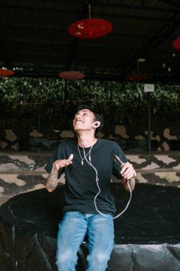 Güneydoğu Asyalı dövmeli adam kulaklıkla müzik dinlemeyi seviyor, rahat ve içerik bir ifadeyle. Boş zaman, kişisel eğlence ya da modern yaşam tarzı temaları için ideal.