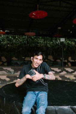 Güneydoğu Asyalı dövmeli adam kulaklıkla müzik dinlemeyi seviyor, rahat ve içerik bir ifadeyle. Boş zaman, kişisel eğlence ya da modern yaşam tarzı temaları için ideal.