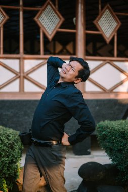 Güneydoğu Asyalı adam belini tutuyor, esniyor ya da rahatlıyor gibi görünüyor. Fiziksel gerginlik, sırt ağrısı veya rahatlama konuları için ideal.