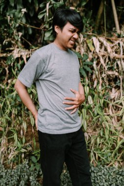 Gergin bir suratı olan Güneydoğu Asyalı bir adam karnını tutarken çok rahatsız oluyor. Acil, rahatsız edici ya da mizahi durumlar için idealdir.