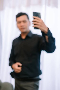 Odaklanamayan bir adamın kapalı bir ortamda telefonunu kullanmasını gösteren soyut bir görüntü. Koyu renk gömlekli adam selfie çekiyor gibi görünüyor. Kasıtlı bulanıklık ise kompozisyona alışılmadık bir çağdaş dokunuş ekliyor. Fotoğraf.