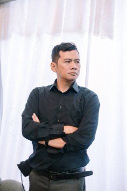 Kolları bağlı bir adam ayakta duruyor. Düşünceli görünüyor ve başka tarafa bakıyor. Koyu renk yakalı bir gömlek giyiyor ve ciddi ama kendine güvenen bir havası var. Liderlik ya da tefekkür için mükemmel bir görüntü..