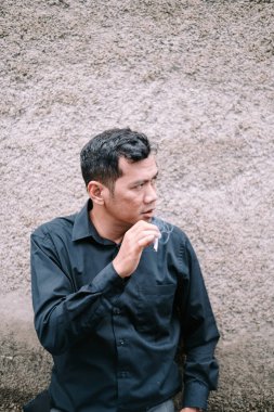 Bir adam dokulu bir duvara yaslanmış sigara içiyor. Düşünceli, düşünceli ya da sorunlu görünüyor. Görüntü bir yansıma ya da tefekkür anını yakalar. Dokuma duvarı tarafsız bir zemin sağlıyor ve dikkatleri bir erkek üzerinde topluyor.