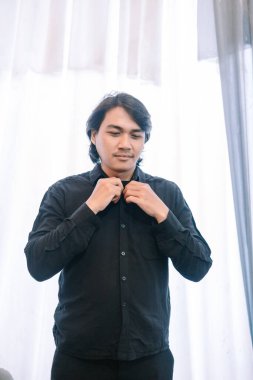 Siyah gömlekli genç bir adam bir hazırlık anında yakasını iliklerken yakalanıyor. Görüntü onun şık kıyafetlerini ön plana çıkarıyor, zarafet ve resmiyet duygusu yaratıyor. Perdelerden süzülen ışık gündüz kuşağını işaret ediyor. 