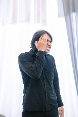 Siyah tişörtlü sıkıntılı bir adam yüzünü eliyle kapatıyor, yüzünü ekşitiyor ve yoğun bir rahatsızlık hissi veriyor, muhtemelen baş ağrısı ya da aşırı stresten. Sahne kapalı, parlak bir arka plan bir pencereyi işaret ediyor.