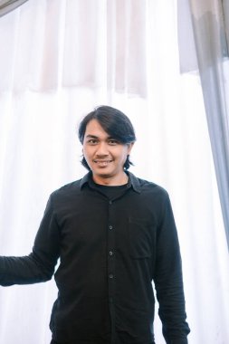 Siyah gömlekli gülümseyen Asyalı bir adam resmediliyor, neşe saçıyor. Basit, parlak zemin onun hoş karşılama ifadesini vurguluyor. Genel duygu modern güveni ve olumlu bir görünümü temsil eder..