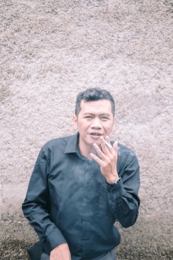 Düşünceli bir adam sigara molası için durur. Resmi bir gömlek giymiş, dokulu bir duvara yaslanmış, dumanı yüzüne doğru kıvrılmış, günlük rutininin ortasında bir anlık düşünceli bir yansıma izlenimi veriyor..