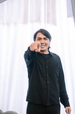 Koyu renk saçlı bir adam güçlü bir bakışla doğrudan izleyiciyi işaret eder. Onun jesti bir aciliyet, suçlama ya da ilgi gerektiriyor. Koyu renk gömleği ve açık arkaplan tarafından oluşturulan katıksız kontrast onun cesur ifadesini vurguluyor. Şey...