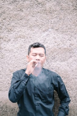 Siyah gömlekli bir adam dokulu bir duvarın önünde duruyor, gözleri kapalı ve parmaklarının arasında bir sigara var. Düşüncelere dalmış gibi görünürken duman havaya karışıyor. Portre, bir yansıma ya da sessiz, kişisel bir anı çağrıştırıyor..