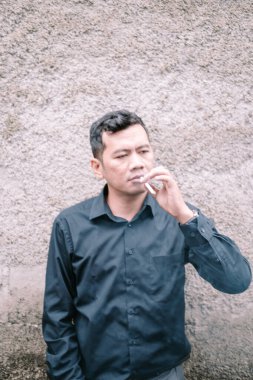 Bir adam, parmaklarının arasında bir sigarayla dokulu bir duvara yaslanır. Koyu renk bir gömlek giyiyor ve derin düşüncelere dalma ya da melankolik bir hava yayıyor. Görüntü samimi bir anı yakalıyor, adamın düşünceli ekspresi arasındaki farkı vurguluyor.