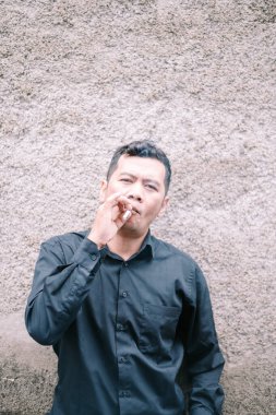 A man dressed in a dark shirt stands against a coarsely textured wall, deeply immersed in thought. A lit cigarette is held between his fingers, emphasizing a moment of introspection and contemplation. The backdrop highlights the subject, focusing on  clipart