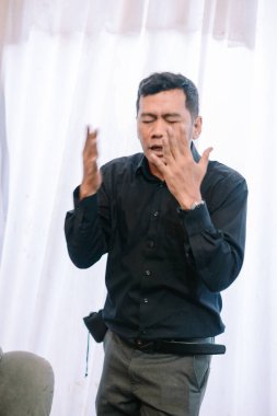 Bir adam güçlü duygular gösterir, ellerini kaldırır ve endişe ya da hüsranla yüzleşir. Koyu renk bir gömlek ve gri pantolon giyiyor, yumuşak beyaz bir perde arkasında duruyor. Görüntü anlamlı bir iletişim anı yakalıyor, muhtemelen despot.