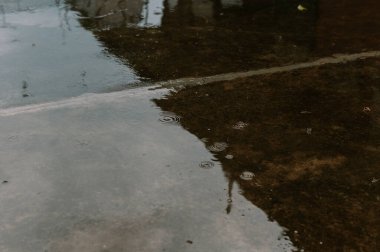 Yağmurun dingin özünü yakalayan minimalist bir kompozisyon. Çembersel dalgalar yağmur damlalarının ıslak betona çarptığı yerden dışarıya doğru yayılarak büyüleyici bir ışık ve gölge etkileşimi yaratır. Kaygan yüzey çevreyi yansıtıyor.