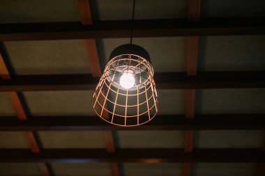 Kafeslenmiş bir ampul, ahşap kirişli bir tavandan sarkmış, yumuşak bir parıltı yayıyor. Sanayi tipi armatür ampulün etrafında bir kablo kafesi içeriyor ve iç uzaya ayırt edici bir tasarım unsuru ekliyor. Tavan ahşap paneller ve b