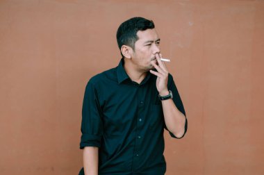 Koyu renk gömlekli bir adam arka planda sigara içmek için duruyor. O düşünceli görünüyor, sanki bu sağlıksız alışkanlığa katılırken derin düşüncelere dalmış gibi. Saat karede görünüyor, biraz da sofistike.