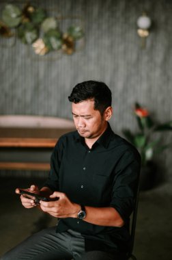 Düşünceli bir adam akıllı telefonuna dalmış, modern tasarım elementlerinin arka planına karşı oturur. Gelişigüzel giyinerek dijital dünyada geziniyor. Yüzündeki ifade odaklanmayı ve bağlanmayı öneriyor. Sahne, sessiz bir bağlantı anını yakalıyor.