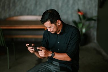 Bir adam cep telefonu cihazıyla etkileşime çok dalmıştır. Rahat bir iç mekan ortamında oturmak. O, sıradan bir düğmeli gömlek giydi. Görüntü, teknolojinin kusursuz entegrasyonuna odaklanmış bir konsantrasyon anı yakalar.