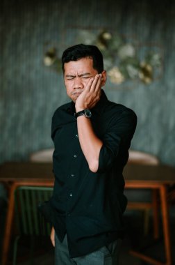 Yüzünde acı dolu bir ifade olan bir adam yüzünü sıkar ve yoğun bir yüz ağrısından muzdarip gibi görünür. Koyu renk gömleği, sıkıntıda kaybolmuş ve yanağını endişeyle tutuyormuş gibi göründüğü için sessiz arka planla çelişiyor. Bu fiziksel bir kopukluk anı yakalar.