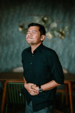 Siyahlar giymiş bir adam midesini tutuyor, kapalı gözlerle rahatsızlık gösteriyor. Mide ağrısı, sindirim sorunları ya da genel hastalık ve mide bulantısı yaşıyor gibi görünüyor. Bu görüntü fiziksel huzursuzluğu vurgular.