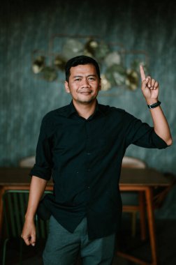 Koyu renk gömlekli kendine güvenen bir adam, yenilikçi bir fikri ya da dikkate alınması gereken önemli bir noktayı sembolize ediyor. Nazik bir gülümsemeyle doğrudan kameraya bakıyor, yaklaşılabilir ve profesyonel bir atmosfer yaratıyor. Ayarlar bir...