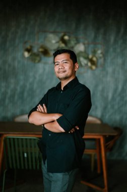 Siyah gömlekli kendine güvenen bir adam kollarını kavuşturup profesyonellik ve özgüven saçıyor. Modern bir ortamda, şık mobilyalar ve ince dekoratif detaylarla geliştirilmiş. Görüntü başarıyı, ulaşılabilirliği ve poziyonu iletir.