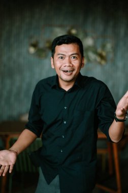 Şaşırmış bir ifadeyle doğrudan kameraya bakan bir adam, ağzı hafifçe açık, dişlerini gösteriyor. Siyah bir gömlek giyiyor ve elleri açık, dışa doğru hareketler yapıyor. Arka plan sessiz, desenli bir duvar. Kaynağa derinlik katıyor.