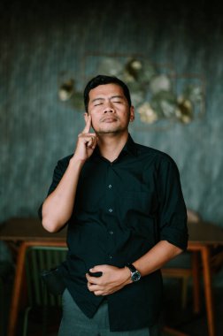 Koyu renk gömlek giyen bir adam gözlerini kapatır, derin bir düşünceye dalıp kaybolur. Parmağıyla şakağına dokunuyor, derin düşüncelere daldığını ve içgözlem yaptığını gösteriyor. Sahne, bir ciddiyet ve odak hissi veriyor, iç dünyayı vurguluyor.