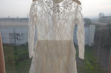 Karmaşık boncuklu ve dantelli zarif bir gelinlik, doğal ışıkla hafifçe aydınlatılmış bir pencereye asılır. Elbisenin tasarımı zarif süsleri içeriyor, incelikli ve romantik bir estetik oluşturuyor. Gelin temalı içerik için ideal ve 