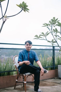 Şık giyinmiş genç bir adam terasta bir sandalyede oturur, düşünceli bir şekilde uzaktan bakar. Sahne, kentsel unsurları doğal yeşilliklerle harmanlayarak sakin ve düşünceli bir atmosfer yaratıyor..