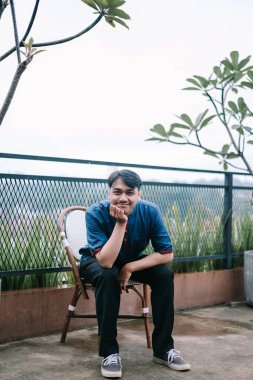 Genç bir Asyalı adam terasta bir sandalyede dalgın dalgın oturuyor. Düşünceli ve rahat görünüyor, şehir manzarasının ortasında huzurlu bir yalnızlığın tadını çıkarıyor. Günlük kıyafetleri ve sakin ifadesi bir memnuniyet hissi veriyor..