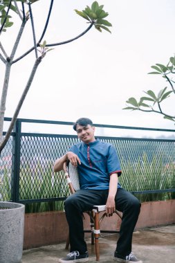 Çatı katındaki terasta rahat rahat oturan gündelik kıyafetli genç bir adam. Nazikçe gülümsüyor ve sakin görünüyor. Şehir manzarasına bakan sakin bir anın tadını çıkarıyor..