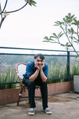 Neşeli genç bir adam dışarıda bir sandalyede oturur, çenesini ellerine koyar ve mutluluk saçar. Huzurlu ortam ve rahat duruş bir memnuniyet ve huzuru çağrıştırıyor..