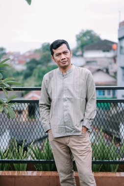 Gündelik giysiler içinde kendine güvenen Güneydoğu Asyalı bir adam şehrin çatısında poz veriyor. Arka planda modern ve şık bir ortam yaratan bir şehir manzarası ve bir çit yer alıyor. Seyahat, yaşam tarzı ya da kültürel temalar için uygun sakin ve huzurlu bir atmosfer yayar.
